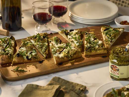 Rao’s pesto flatbread topped with melted fresh mozzarella, arugula, and balsamic glaze, sliced and served on a wooden board.