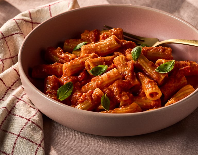Bowl of rigatoni pasta with Rao's® tomato sauce and fresh basil on a rustic table setting